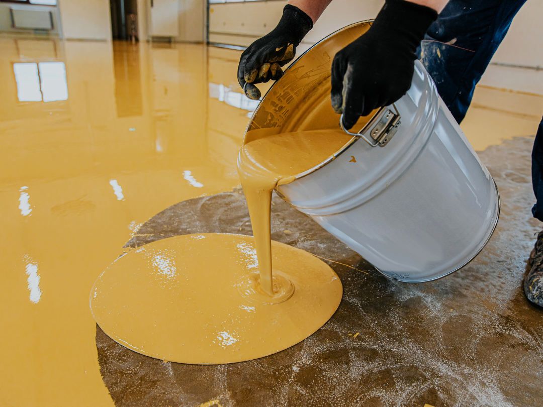 Worker pouring and spreading yellow epoxy mixture evenly on the floor of a commercial area.